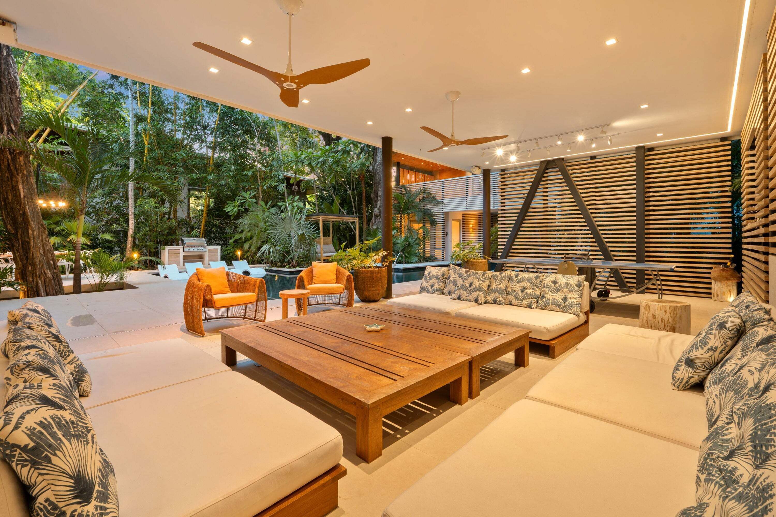 A modern outdoor lounge area with a wooden table, sofas, and chairs, surrounded by lush greenery and a pool.