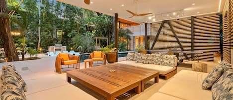 A modern outdoor lounge area with a wooden table, sofas, and chairs, surrounded by lush greenery and a pool.