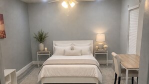 Quarto com cama, mesas de cabeceira, escrivaninha, cadeira, ventilador de teto e janela com persianas.