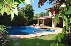Piscina com deck de sol, cercada por vegetação exuberante e um edifício de dois andares com varanda coberta.