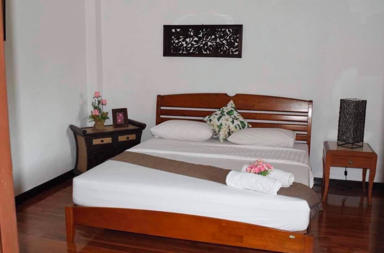 Un dormitorio con cama de madera, ropa de cama blanca, una mesita de noche con un florero y un panel decorativo en la pared.