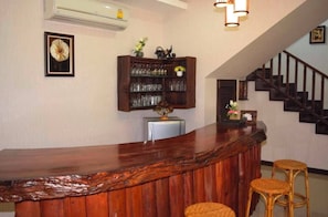 Uma área de bar com balcão de madeira, banquetas de vime, armário para copos e uma escada ao fundo.