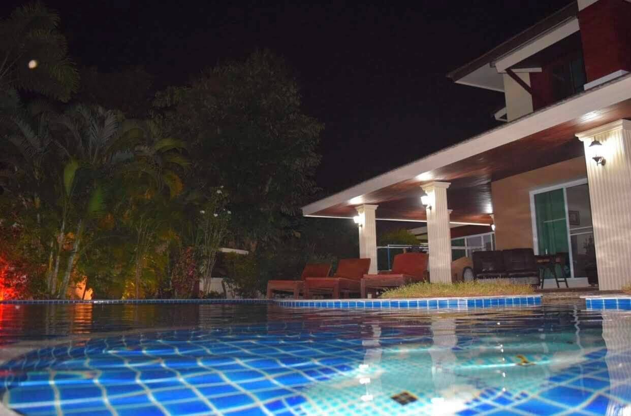 Une piscine dont le bord est revêtu de carreaux, entourée de palmiers et bordée d’un bâtiment doté d’un patio couvert.