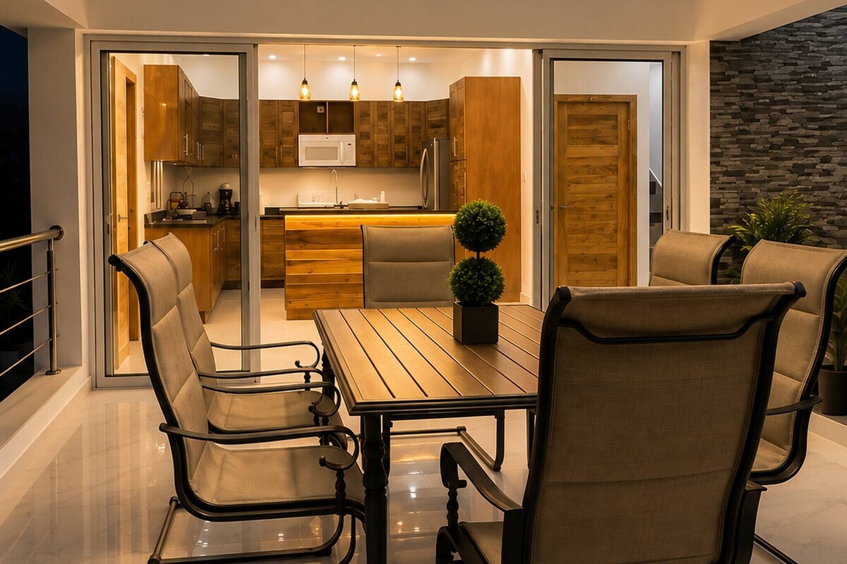 A modern kitchen with a dining area, featuring a wooden table and chairs, a ceiling fan, and a view into the adjacent living space.