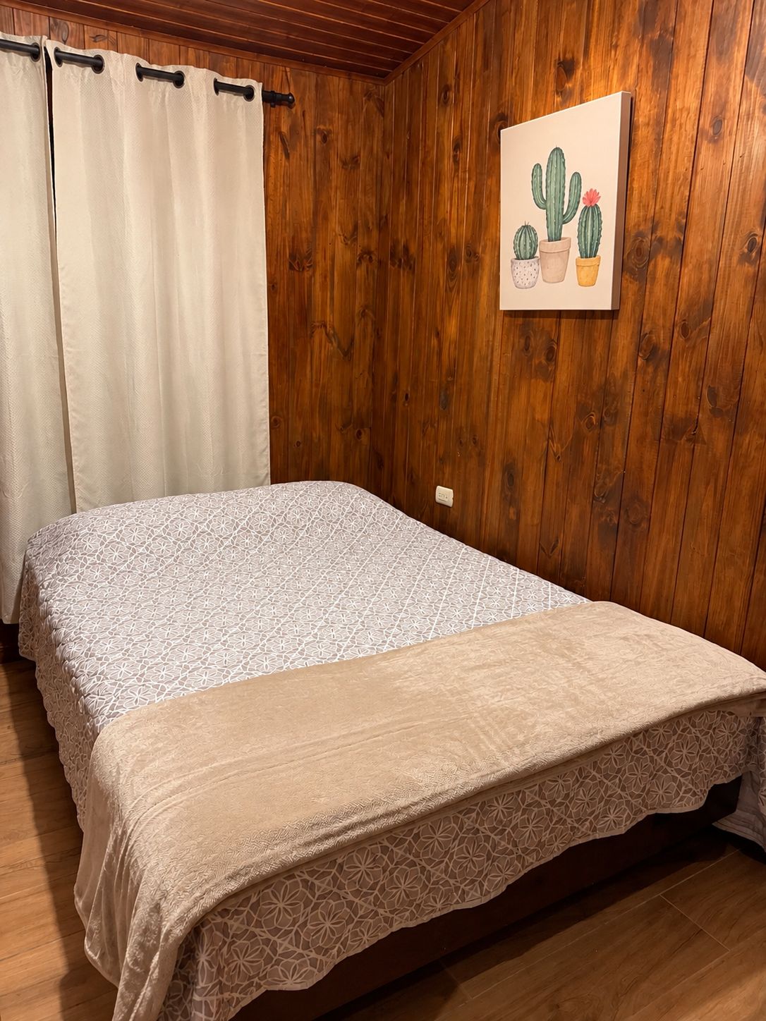 A wooden-paneled room with a bed, curtains, and a wall-mounted painting of cacti.