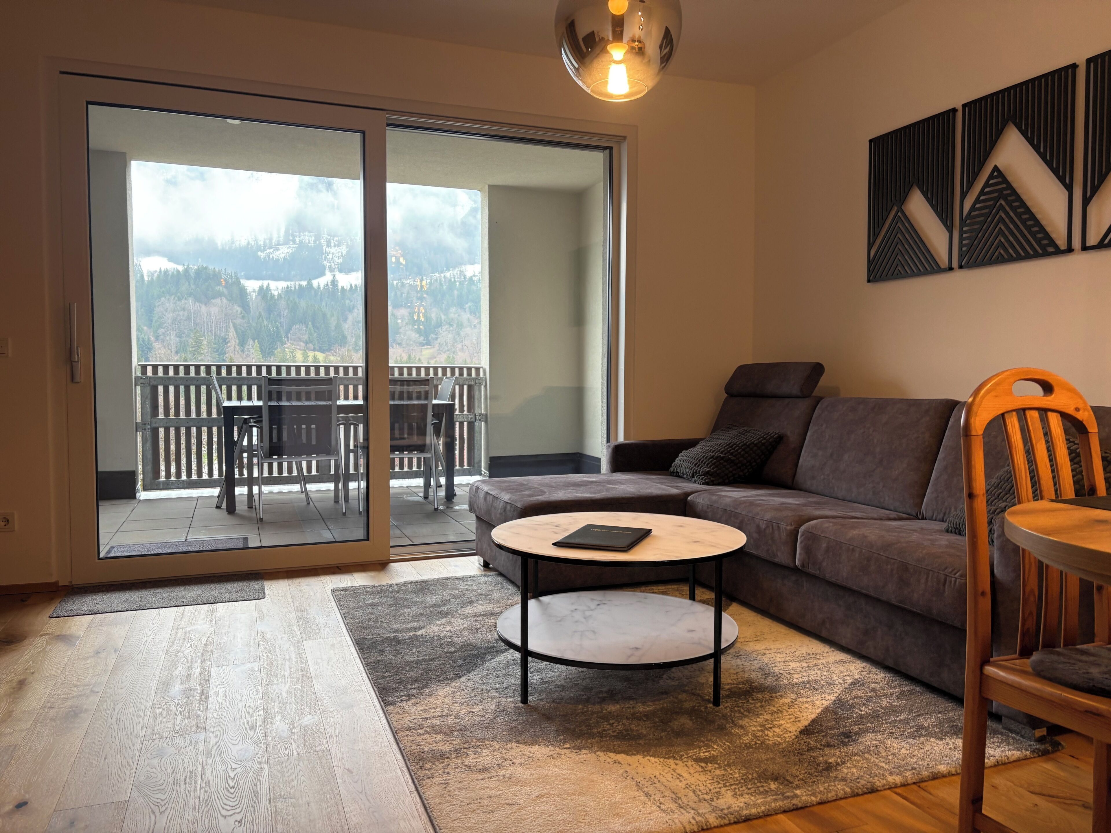 Una sala de estar moderna con un sofá gris, una mesa de centro redonda y una silla de madera. Se ve un balcón con vistas a las montañas a través de puertas de vidrio.