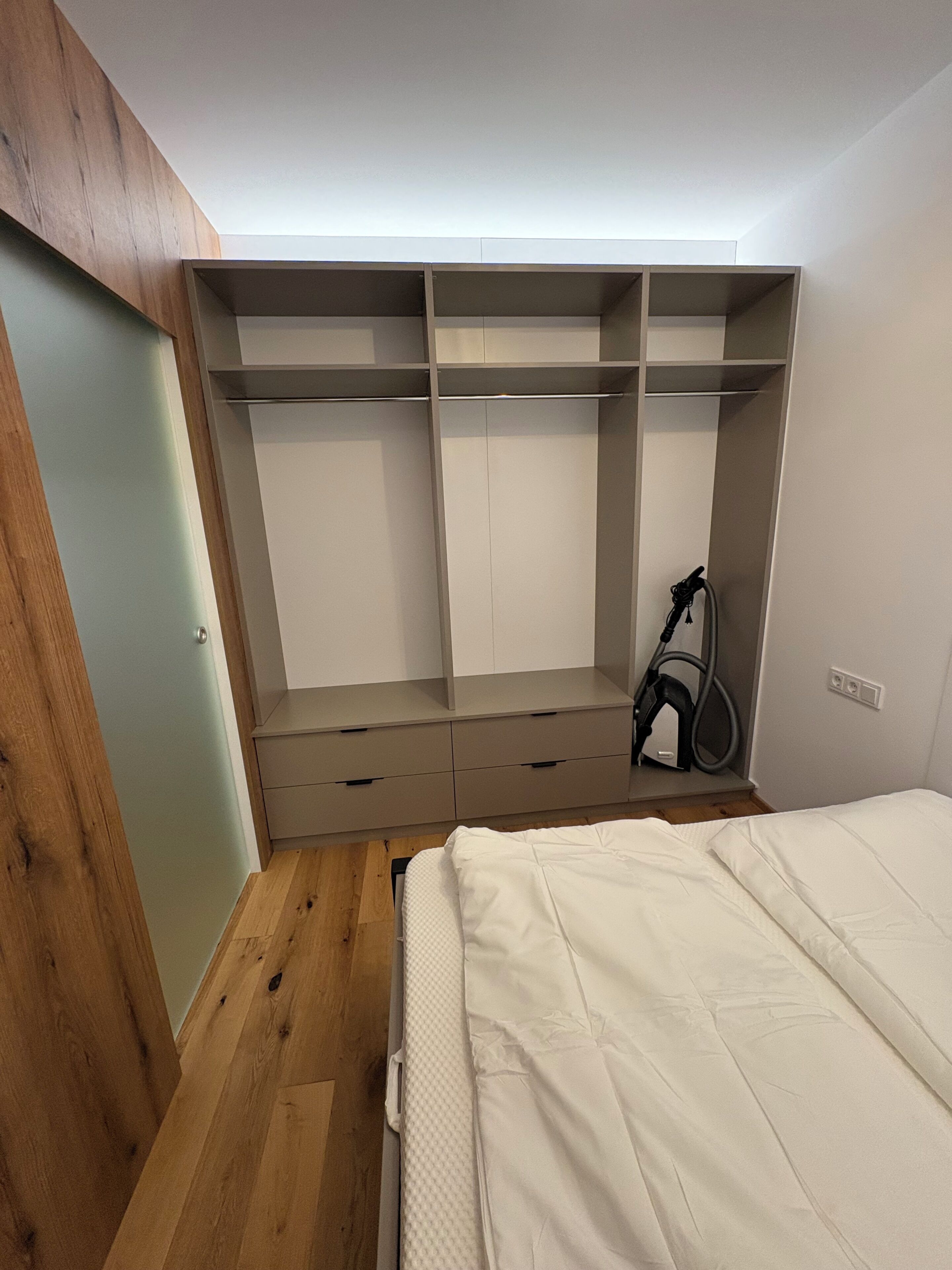 Une chambre à coucher avec un lit, un plancher en bois, une armoire intégrée dotée de plusieurs compartiments et un aspirateur.