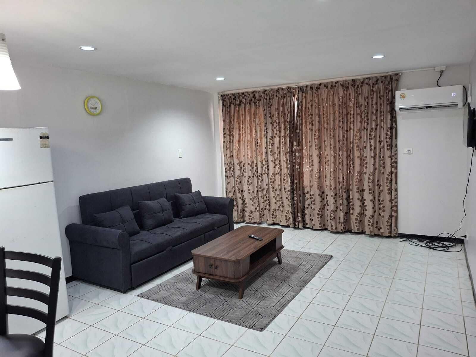 Una sala de estar con un sofá oscuro, una mesa de centro de madera, un tapete estampado, un refrigerador blanco y una ventana con cortinas florales.