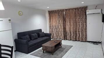 Una sala de estar con un sofá oscuro, una mesa de centro de madera, un tapete estampado, un refrigerador blanco y una ventana con cortinas florales.