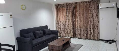 A living room with a dark sofa, a wooden coffee table, a patterned rug, a white refrigerator, and a window with floral curtains.
