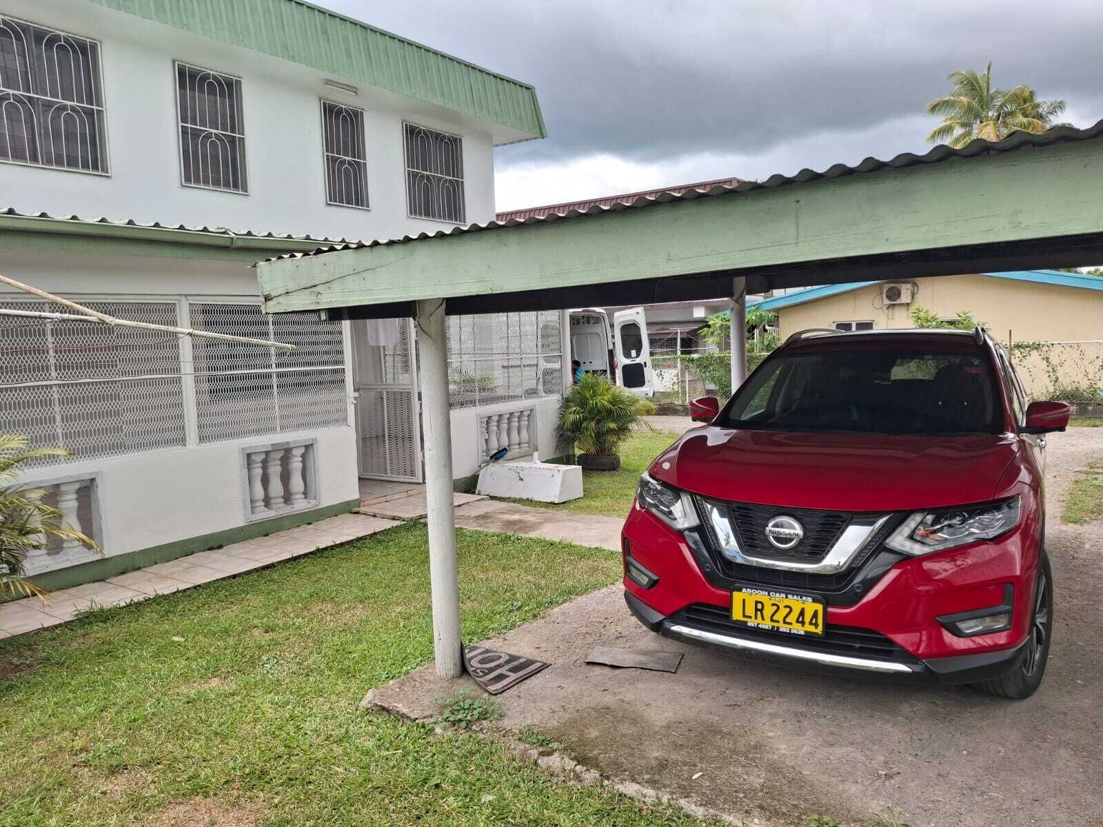 Un Nissan SUV rojo estacionado bajo una estructura cubierta, con un edificio blanco al fondo.