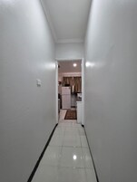 Un pasillo estrecho con paredes blancas y suelo de baldosas que conduce a una zona de cocina con un refrigerador y un microondas.