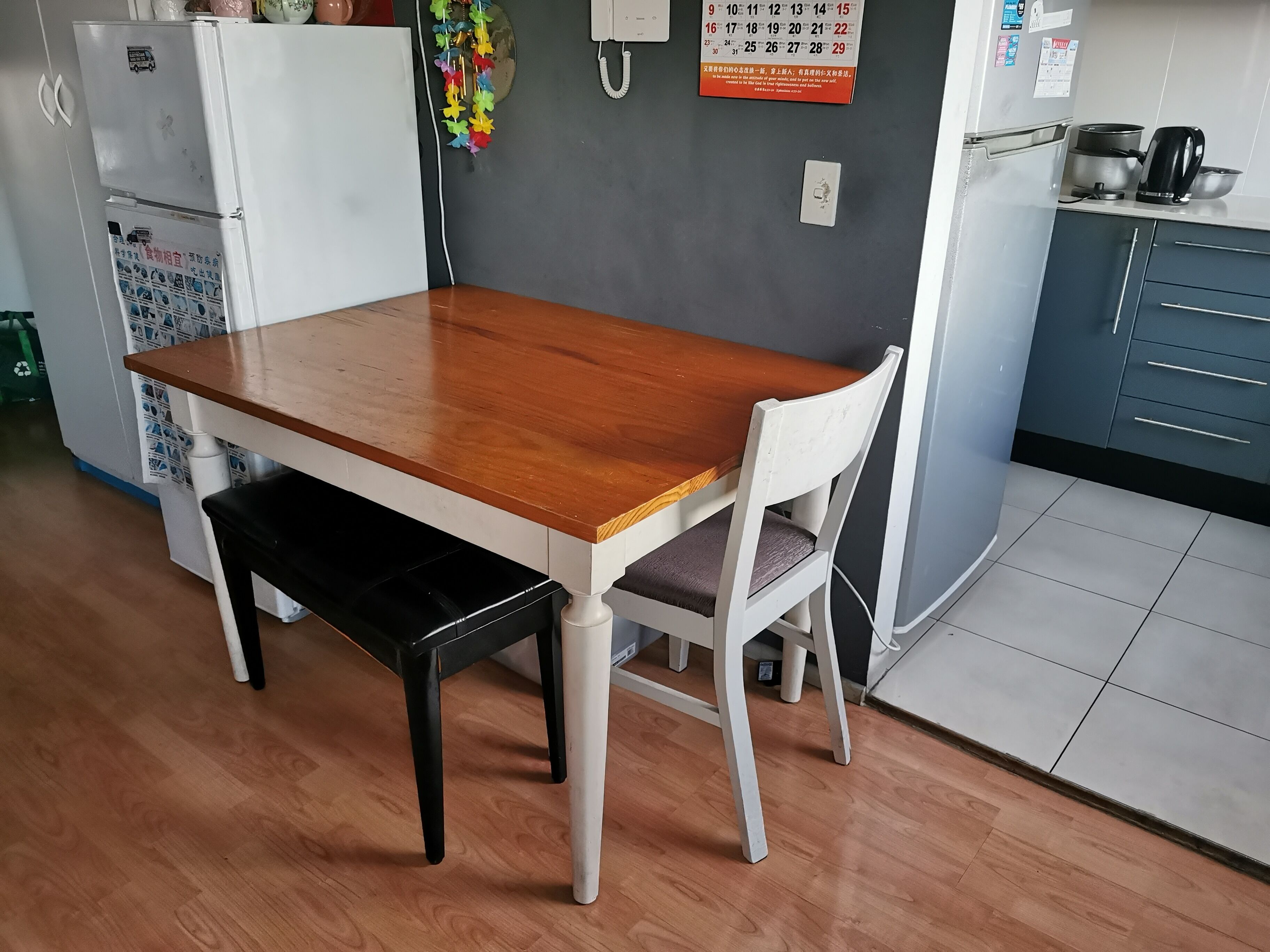 A dining area with a wooden table and chairs, a refrigerator, a kettle, and a calendar on the wall.