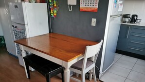 Ruokailutilassa on puinen pöytä ja tuolit, jääkaappi, vedenkeitin ja seinällä kalenteri.