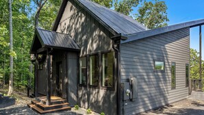 A small, dark-colored cabin with a steep roof, a porch, and a small deck. - Luxe Cabin with Heated Indoor Pool, Hot Tub, Arcade & Theater near Dollywood (Sevierville)