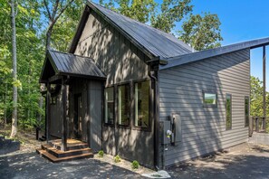 A small, dark-colored cabin with a steep roof, a porch, and a small deck. - Luxe Cabin with Heated Indoor Pool, Hot Tub, Arcade & Theater near Dollywood (Sevierville)