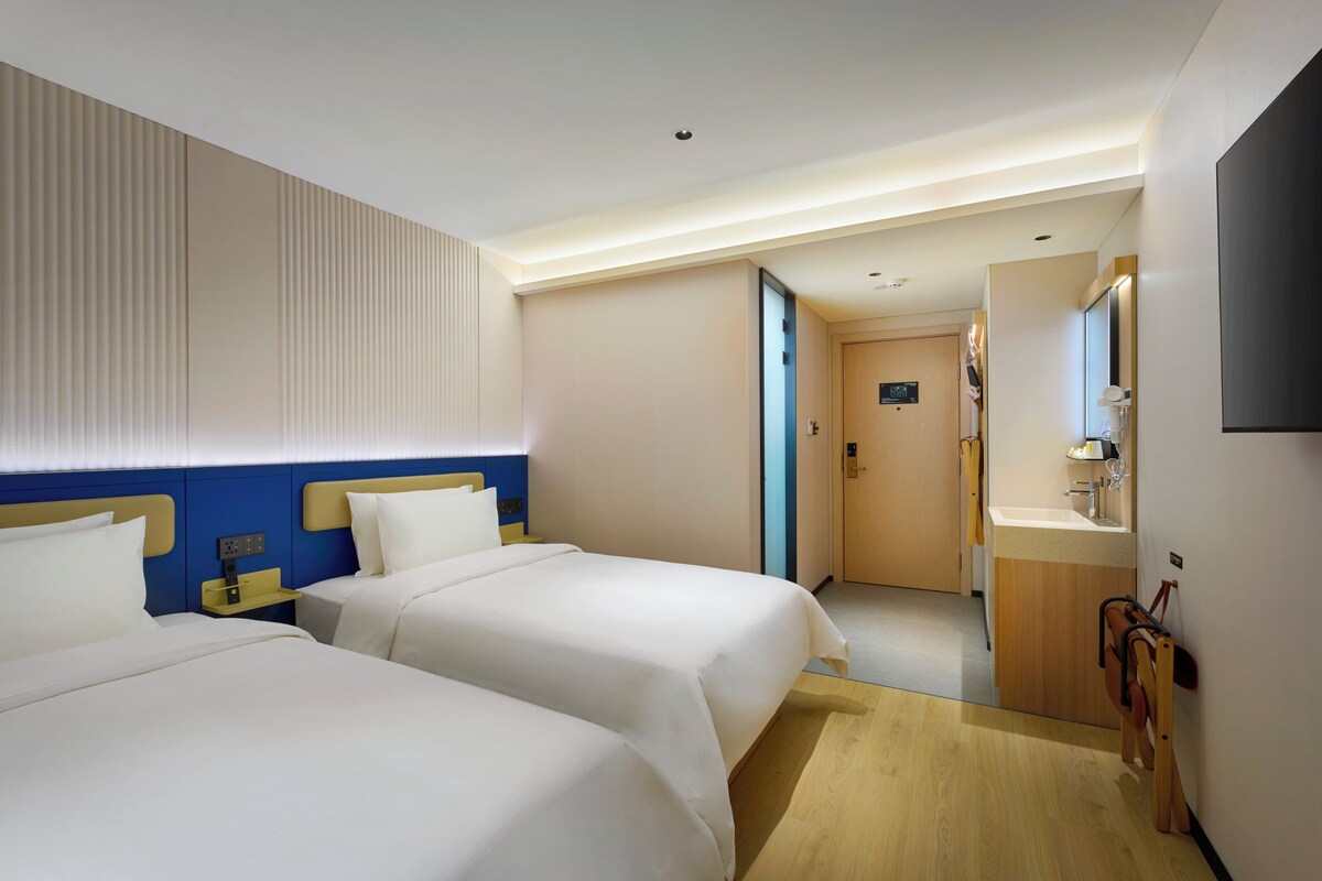 Habitación de hotel con dos camas, piso de madera, baño con lavamanos y espejo, y una puerta que da a otra habitación.