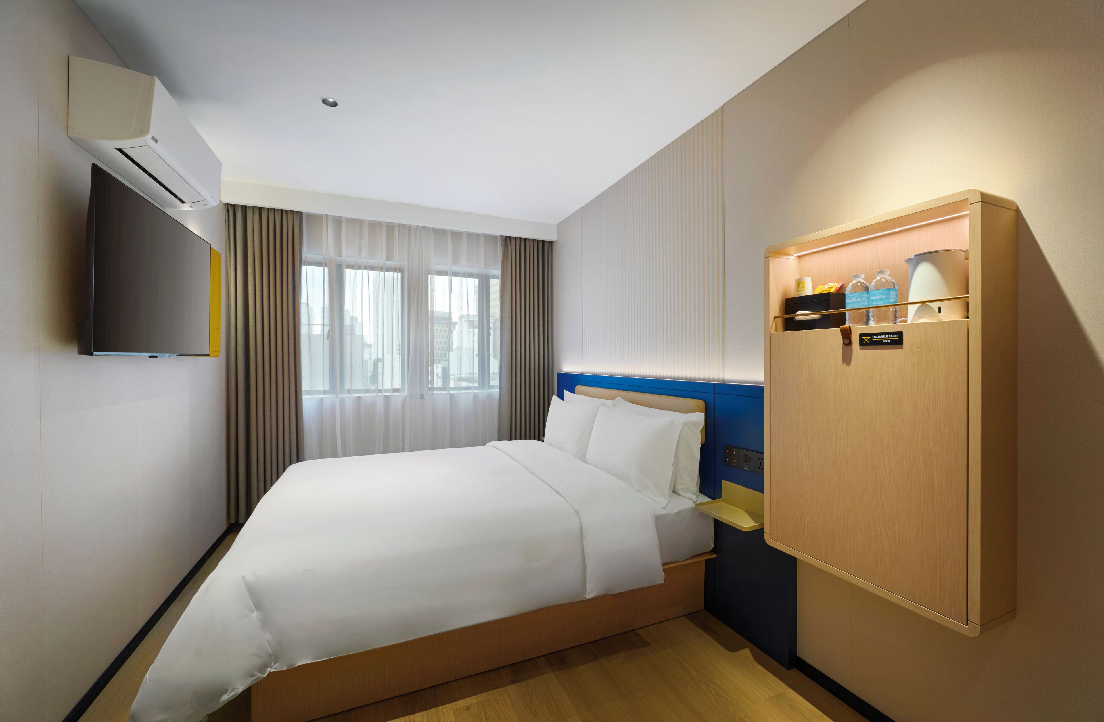 Habitación de hotel con cama, televisor montado en la pared, ventana con cortinas y armario de madera con etiqueta.
