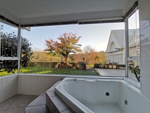 A modern bathroom with a large bathtub, a view of a garden with trees and a lawn, and a white tiled floor.
