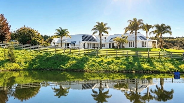 Um edifício branco espaçoso, com várias janelas e painéis solares, rodeado por vegetação exuberante e palmeiras, com um corpo de água calmo ao primeiro plano.
