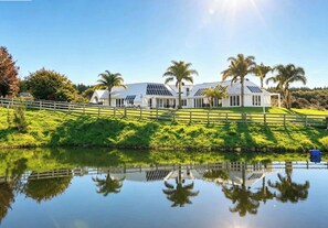 A large white building with multiple windows and solar panels, surrounded by lush greenery and palm trees, with a calm body of water in the foreground.
