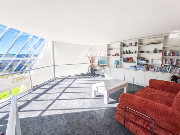 A modern living room with a large window, a red sofa, a white coffee table, and a bookshelf filled with books and decorative items.