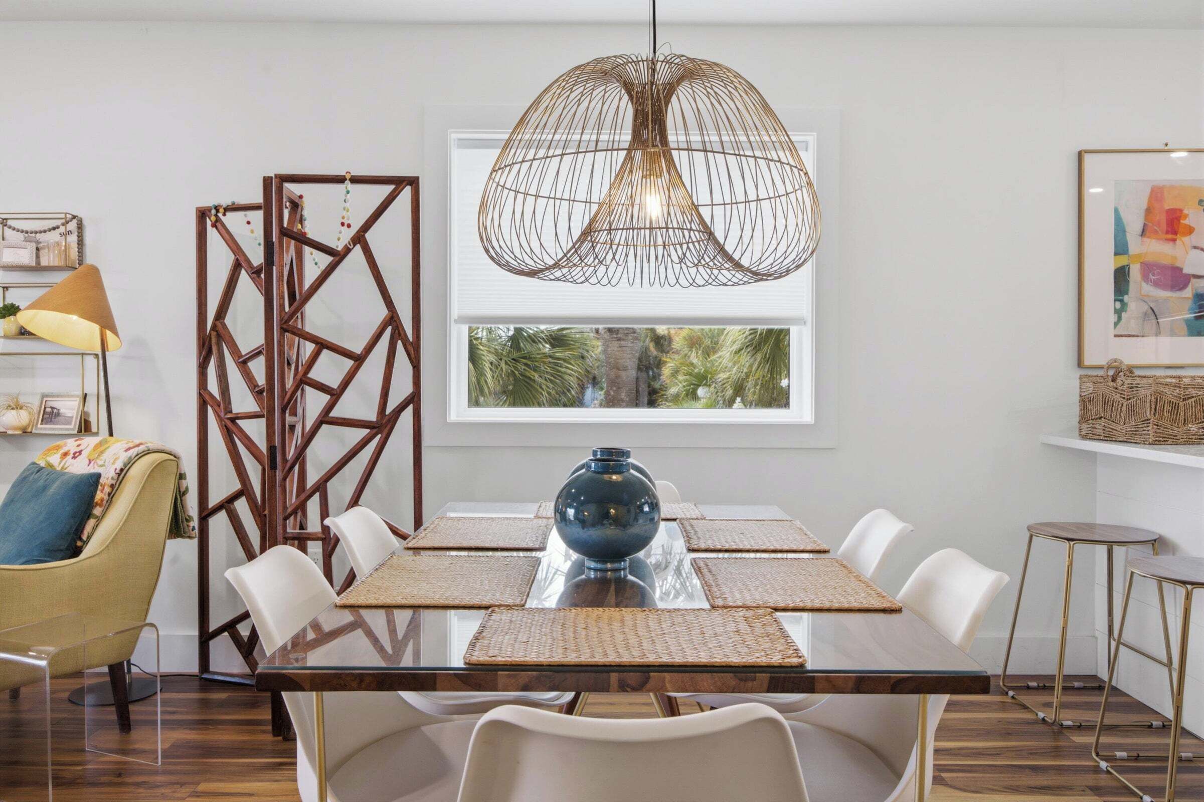 A dining area with a glass table, white chairs, a blue vase, a geometric wooden partition, and a hanging light fixture.
