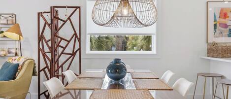 Un comedor con una mesa de vidrio, sillas blancas, un jarrón azul, un separador geométrico de madera y una lámpara colgante.