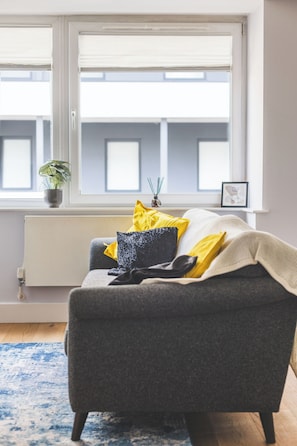 A modern living room with a grey sofa, yellow and black cushions, a blue patterned rug, a potted plant, and a radiator. - Stylish 2BR | Fast Links to Central London (Croydon)