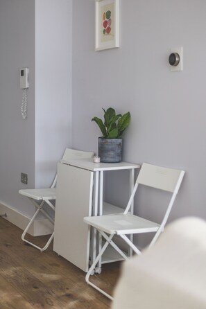 A room with a white foldable desk, a white chair, a potted plant, a wall-mounted phone, and a doorbell. - Stylish 2BR | Fast Links to Central London (Croydon)