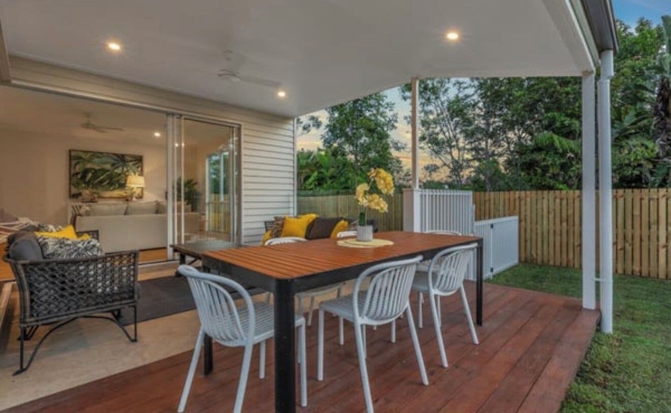 Une terrasse extérieure avec une table en bois et des chaises blanches, un espace salon abrité et une vue sur le jardin arrière.