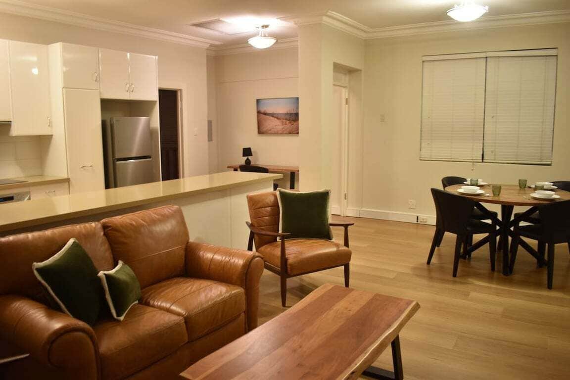 A modern kitchen with a built-in refrigerator, a dining area with a wooden table and chairs, and a leather sofa with cushions.