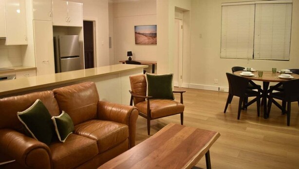 A modern kitchen with a built-in refrigerator, a dining area with a wooden table and chairs, and a leather sofa with cushions. - City Loft | Stylish CBD Stay + River Walks (Rockhampton)