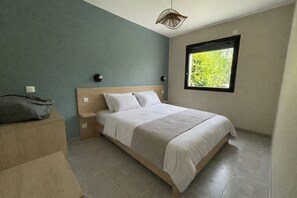 A bedroom with a bed, nightstands, a dresser, a window with a view of greenery, and a pendant light. - Vrbo Property (La Plaine-sur-Mer)