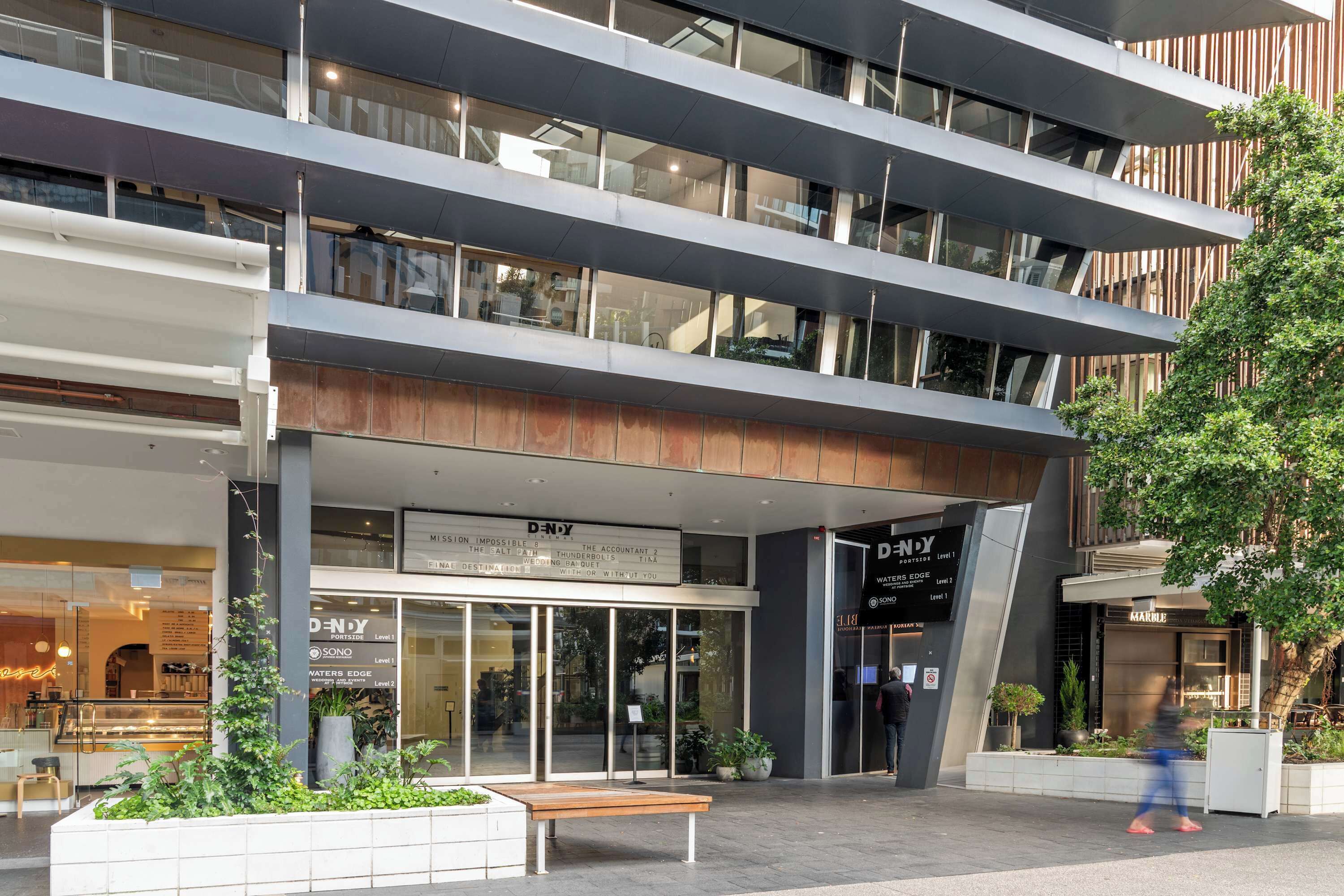 A multi-story building with a modern architectural design, featuring a ground-level entrance with glass doors, signage for a cinema, and a seating area with a bench.