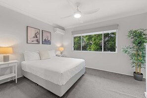 Ein Schlafzimmer mit einem Bett, zwei Nachttischen, einem Deckenventilator, einem Fenster mit Blick auf Grünflächen und einer Topfpflanze.