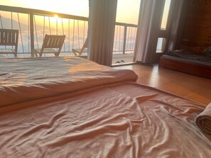 A bedroom with a bed, a wooden floor, a balcony with chairs, and a view of the sky.