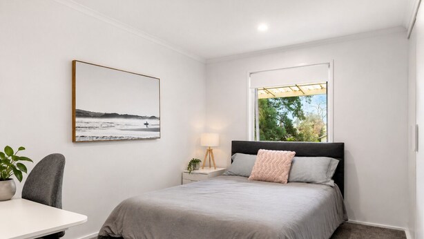 Quarto com cama, escrivaninha, cadeira, mesa de cabeceira com abajur, janela com vista para árvores e um quadro na parede.