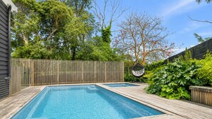 Piscina retangular com um deck de madeira ao redor, cerca de madeira e vegetação exuberante ao fundo.