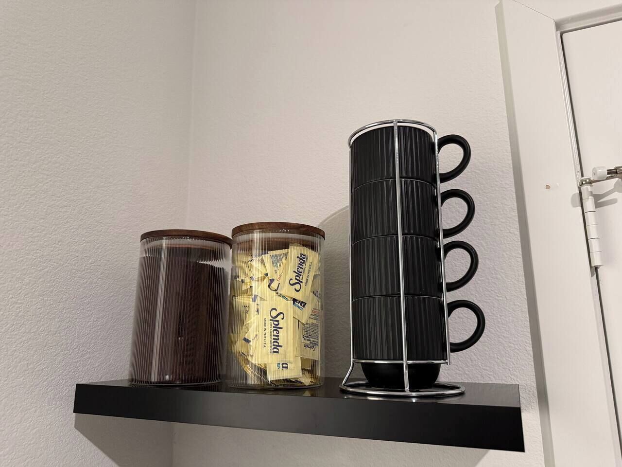 A shelf with three containers: a clear glass jar, a clear plastic jar with a label, and a black cylindrical container with rings.