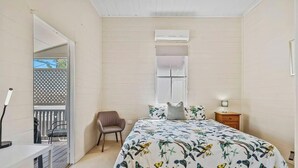 A bedroom with a bed, a chair, a nightstand, an air conditioning unit, and a window. - City Cottage (Rockhampton)