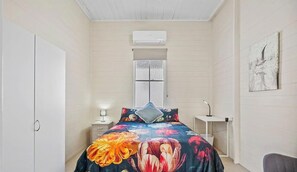 A bedroom with a bed, a nightstand, a desk, a chair, a wardrobe, and an air conditioning unit.