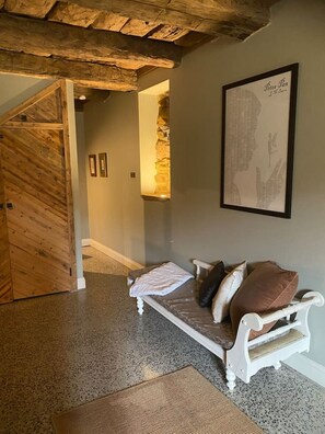 Una habitación con puerta de madera, un banco con cojines, un cuadro enmarcado en la pared y una alfombra con textura en el suelo.