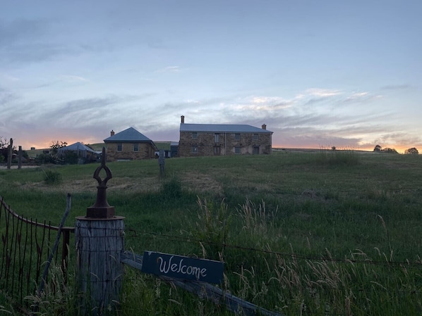 A rural landscape with a stone building and a wooden fence with a 'Welcome' sign.