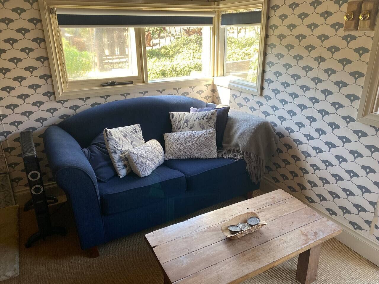 A room with a blue sofa, patterned wallpaper, a wooden table, and a window with a view of greenery.
