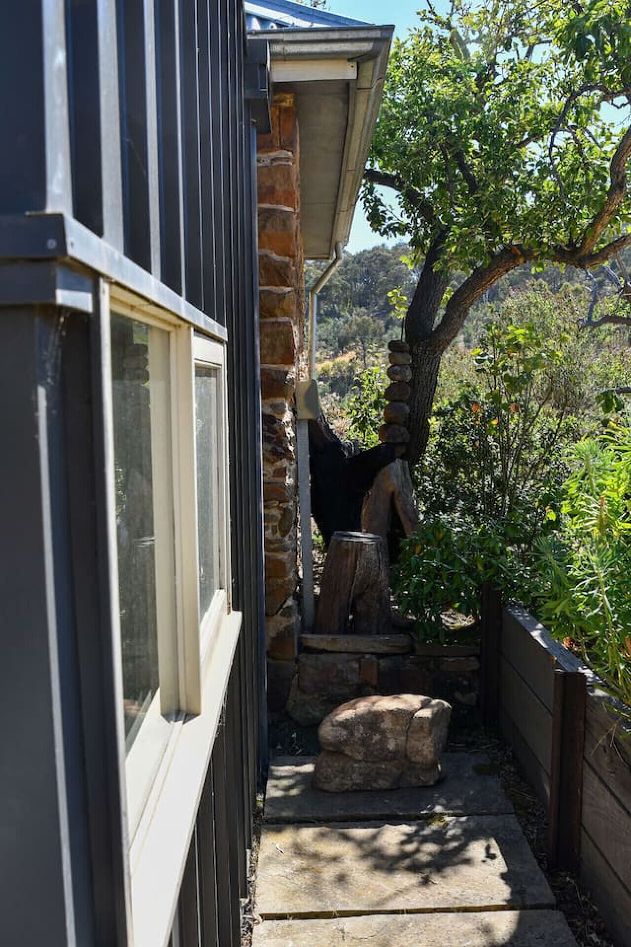 A narrow outdoor pathway leading to a stone bench, with a stone wall and a tree on one side and a building with a window on the other.