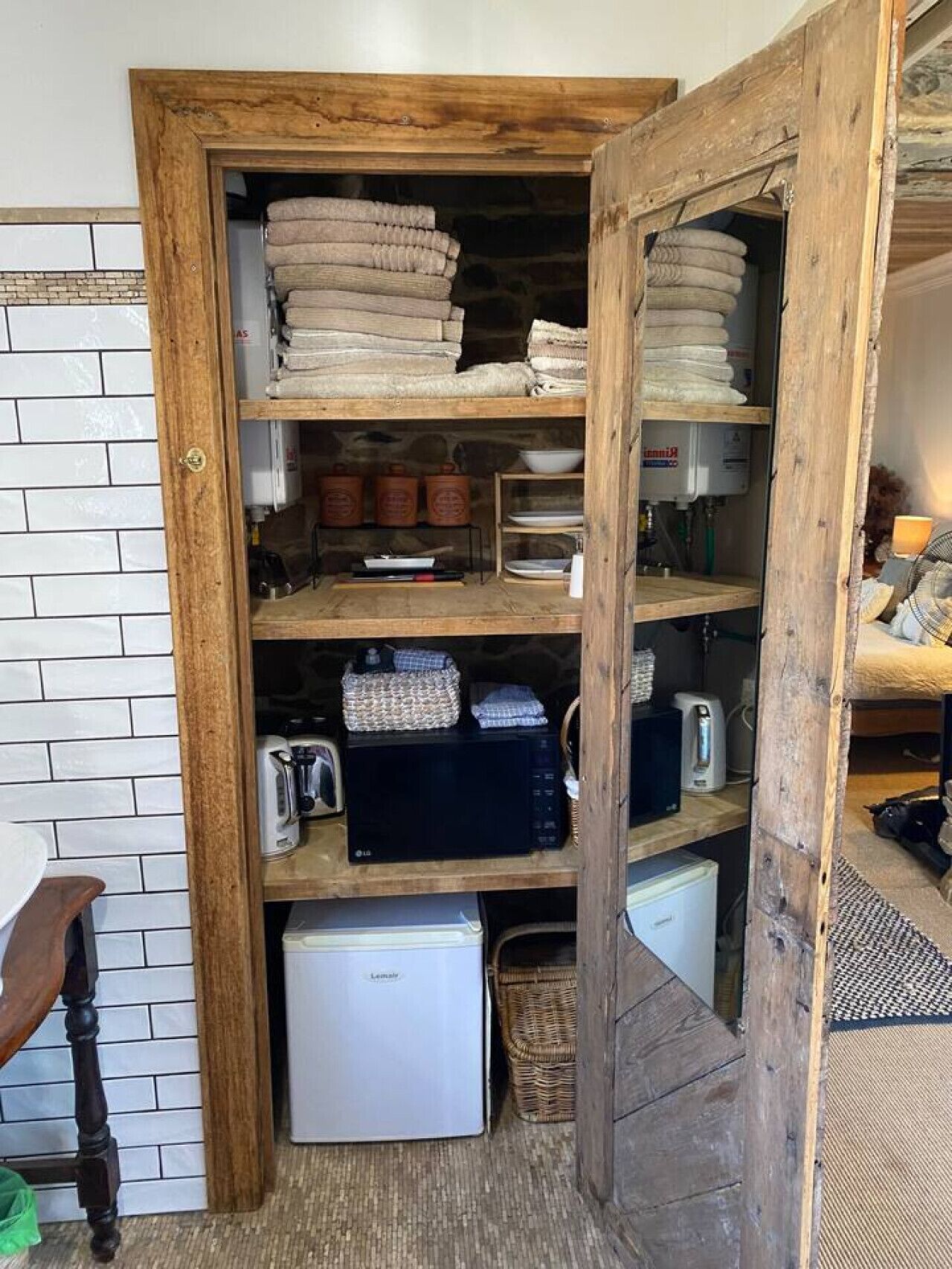 A compact storage area with shelves containing folded towels, kitchenware, and a small refrigerator.