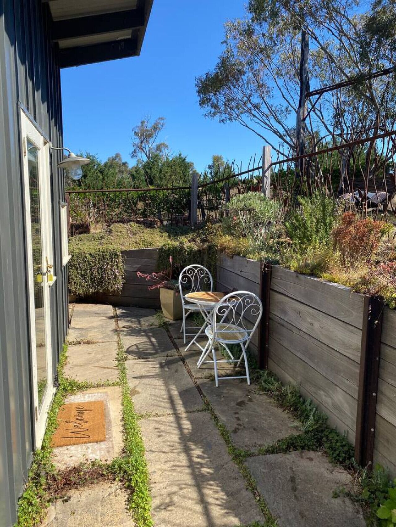 A patio with a metal table and chairs, a wooden fence, and a concrete pathway leading to a door.