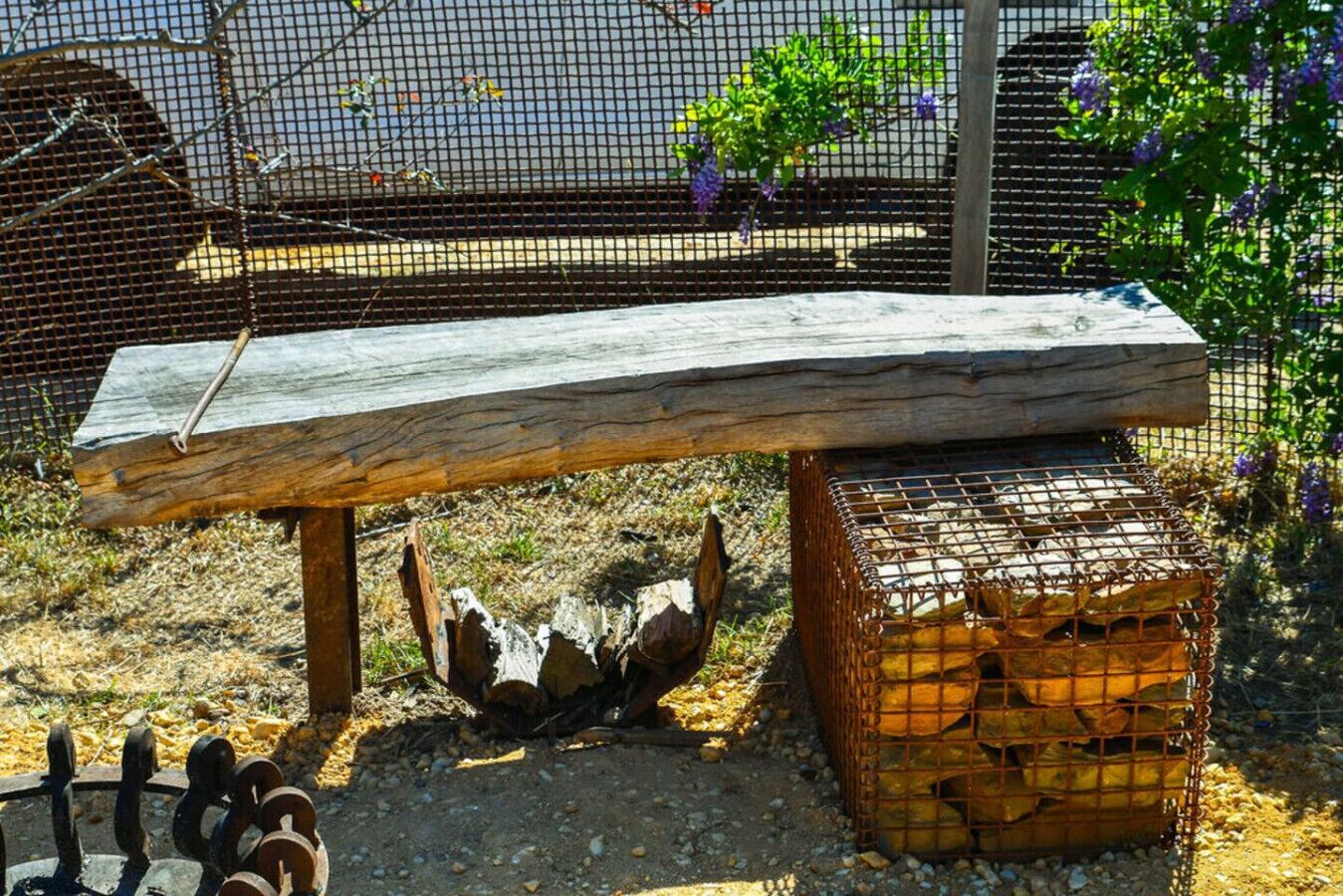 A rustic outdoor setting with a wooden bench, a metal cage, and a pile of logs.