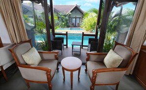 A room with two wooden chairs and a small table, a glass door leading to a pool area with a view of a house and palm trees. - Gokulam Grand Resort and Spa Kumarakom (Kottayam)